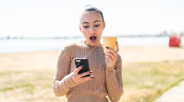 young moroccan girl outdoors using mobile phone holding coffee with surprised expression 1368 466533